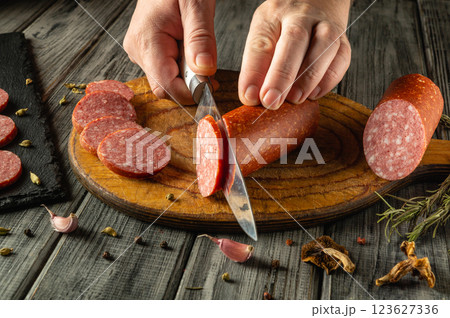 Hands skillfully cut slices of salami on a textured wooden board. Surrounding ingredients include garlic cloves and herbs, creating a warm culinary atmosphere Hands skillfully cut slices of salami on a textured wooden board. Surrounding ingredients include garlic cloves and herbs, creating a warm culinary atmosphere 123627336