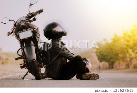 Motorcyclist sitting next to his motorcycle on the road. Motorcyclist sitting and leaning on his motorcycle on the asphalt Motorcyclist sitting next to his motorcycle on the road. Motorcyclist sitting and leaning on his motorcycle on the asphalt 123627619