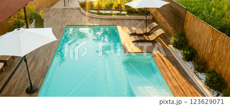 Modern swimming pool with turquoise water, wooden deck, umbrellas, and lounge chairs in a tranquil backyard Modern swimming pool with turquoise water, wooden deck, umbrellas, and lounge chairs in a tranquil backyard 123629071