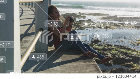 Social media notifications image over woman sitting by ocean taking photos 123634414