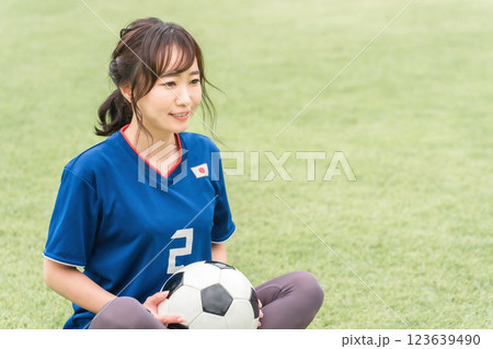 サッカーのユニフォーム姿でサッカーボールを持って芝生グランドの上に座る女性 123639490