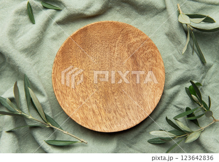 Empty round wooden plate on green linen fabric with olive branches decoration Empty round wooden plate on green linen fabric with olive branches decoration 123642836