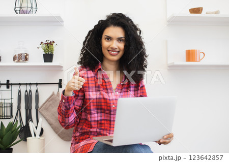 African young woman in casual outfit sitting on countertop in kitchen holding laptop smiling giving thumbs up symbolizing positivity, successful online work, demonstrating happiness. 123642857