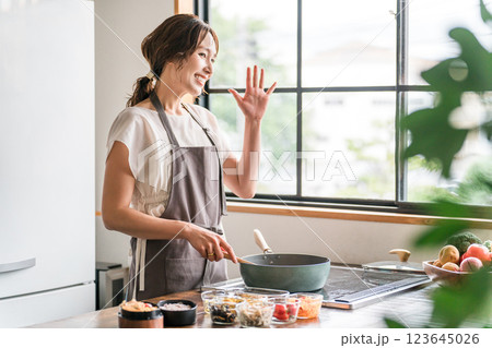 キッチンでフライパンを使うエプロン姿の女性（笑顔・手を振る） 123645026