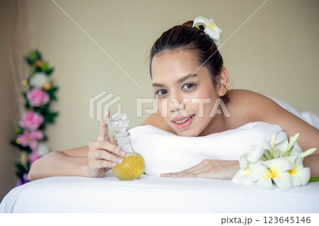 Smiling woman lying on massage bed in spa, holding glass bottle of essential oil while surrounded by white plumeria flowers, enjoying relaxing aromatherapy session for beauty and wellness 123645146