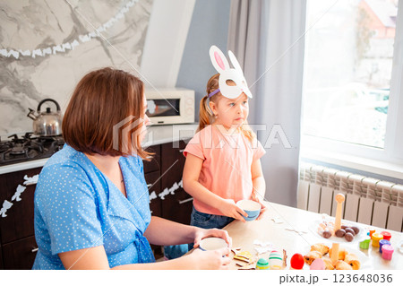 Wide shot of Caucasian mother and her little daughter are drinking tea together in the kitchen. Little girl put on a homemade paper rabbit mask. Concept of the Easter Spring festival Wide shot of Caucasian mother and her little daughter are drinking tea together in the kitchen. Little girl put on a homemade paper rabbit mask. Concept of the Easter Spring festival 123648036
