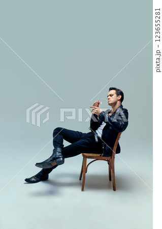 Man dressed vintage attire lounging effortlessly, cigarette lit with timeless confidence, embodying essence of cinematic cool against studio background. Man dressed vintage attire lounging effortlessly, cigarette lit with timeless confidence, embodying essence of cinematic cool against studio background. 123655281