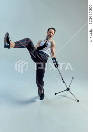 Young stylish man, singer kicking in expressive rhythm while gripping microphone, fusing dance and music into one vibrant moment against studio background. Young stylish man, singer kicking in expressive rhythm while gripping microphone, fusing dance and music into one vibrant moment against studio background. 123655306
