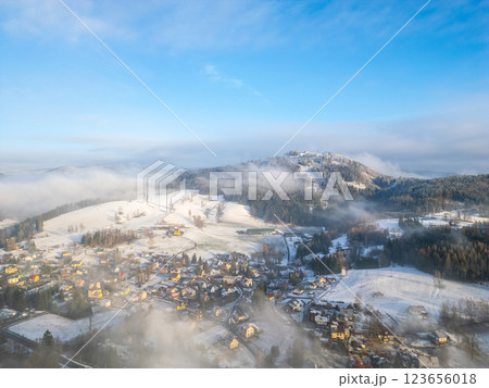Snow blankets the hills and houses around Tanvaldsky Spicak, creating a peaceful winter vista. Low clouds drift among the trees, enhancing the serene atmosphere of the landscape. 123656018