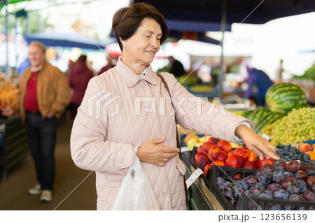 Positive mature woman in casual clothes picking fresh plums during shopping at grocery market Positive mature woman in casual clothes picking fresh plums during shopping at grocery market 123656139