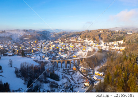 A stone railway bridge spans over a snowy valley in Smrzovka, Czechia. The winter landscape features frosted trees and quaint houses, creating a picturesque scene in a serene atmosphere. A stone railway bridge spans over a snowy valley in Smrzovka, Czechia. The winter landscape features frosted trees and quaint houses, creating a picturesque scene in a serene atmosphere. 123656585