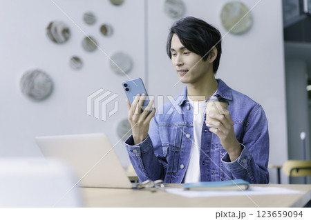 スマホとコーヒーを手にpc作業をする若い男性　撮影協力:スカパー東京メディアセンター 123659094