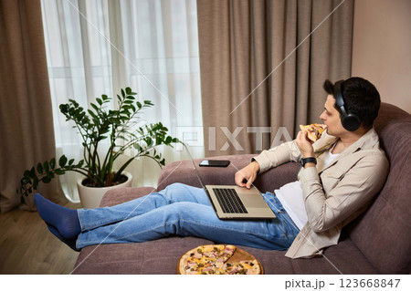 Young man sitting on sofa and eating pizza while watching movie on laptop 123668847