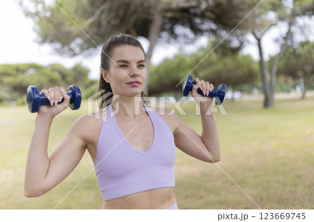 A Vibrant Young Woman Enthusiastically Lifting Weights in the Park on a Beautiful, Sunny Day 123669745