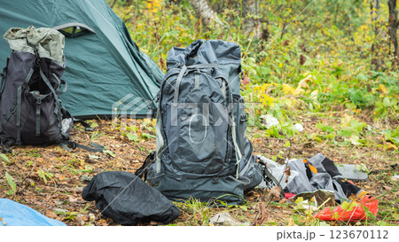 A large gray hiking backpack stands in the foreground, next to a partially visible green tent. Other hiking gear is scattered around, including the backpack and rain cover. Mountain Hiking A large gray hiking backpack stands in the foreground, next to a partially visible green tent. Other hiking gear is scattered around, including the backpack and rain cover. Mountain Hiking 123670112
