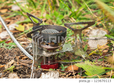 A portable camping stove with a metal burner and a support frame stands on a bed of fallen leaves and needles in the forest. A gas cylinder is connected to the stove via a hose. Camping equipment A portable camping stove with a metal burner and a support frame stands on a bed of fallen leaves and needles in the forest. A gas cylinder is connected to the stove via a hose. Camping equipment 123670113