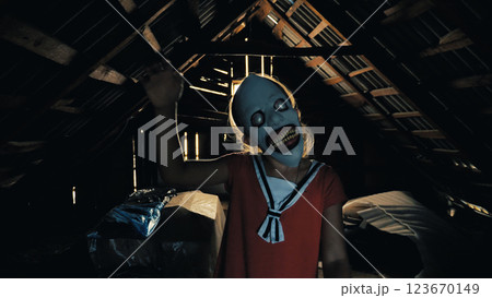 A young girl wearing a creepy mask with big eyes and a wide smile stands in a dark, shadow-covered attic. She waves her right hand. Close-up of a child monster A young girl wearing a creepy mask with big eyes and a wide smile stands in a dark, shadow-covered attic. She waves her right hand. Close-up of a child monster 123670149