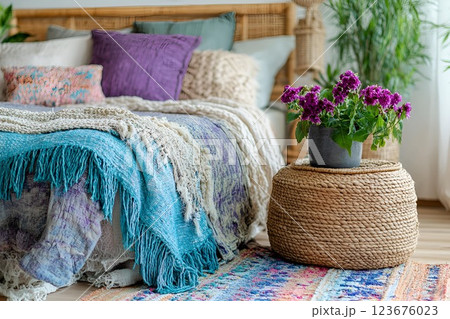 Cozy bohemian bedroom with a bed covered in textured blankets, decorative pillows, a woven ottoman, and a potted flower 123676023
