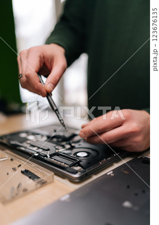 Vertical cropped shot of electronics repair professional carefully disassembling laptop motherboard, precision screwdriver in hand, surrounded by various components on workshop table, close-up. 123676135