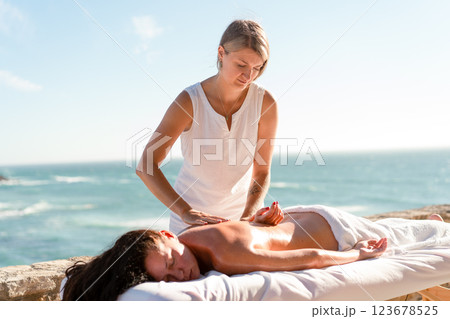 Therapist massaging woman's back on table by sea Therapist massaging woman's back on table by sea 123678525