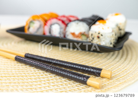 Variety of sushi rolls on black plate with chopsticks on woven mat Variety of sushi rolls on black plate with chopsticks on woven mat 123679789