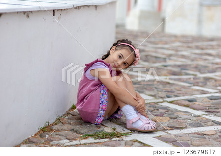 Young hispanic girl sitting on cobblestone street wearing pink dress and headband 123679817