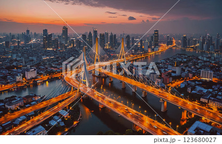 City at night with a bridge and a river. The city is lit up with lights and the sky is orange 123680027