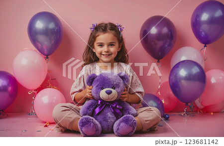 Young girl is sitting on the floor holding a purple teddy bear. The room is decorated with many balloons, creating a festive atmosphere. The girl is smiling and he is enjoying her time with the teddy 123681142