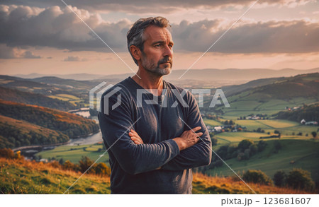 Man is standing on a hill with his arms crossed, looking out over a vast, green landscape 123681607