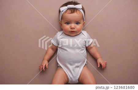Baby wearing a white outfit and a bow in her hair. The baby is laying on a brown surface 123681680