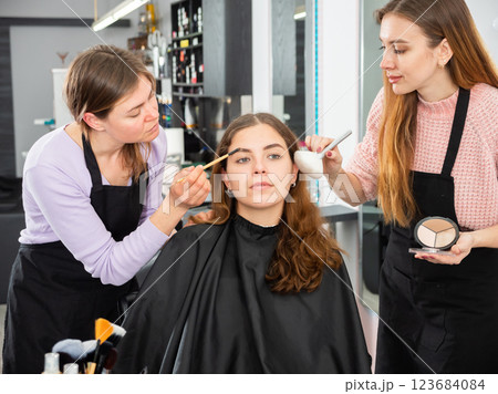 Two makeup artists apply makeup on the face of a young woman 123684084