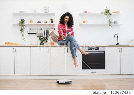 Young African woman in modern kitchen sitting on countertop using smartphone observing robotic vacuum cleaner performing tasks on floor Young African woman in modern kitchen sitting on countertop using smartphone observing robotic vacuum cleaner performing tasks on floor 123687420