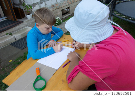 Young boy and older woman drawing together outside at a wooden table, with paper, ruler, pens, and a magnifier. Young boy and older woman drawing together outside at a wooden table, with paper, ruler, pens, and a magnifier. 123692591