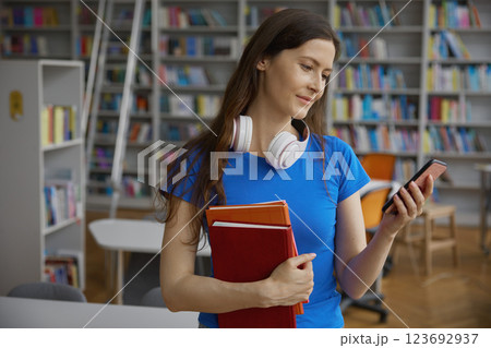 Young woman reading email message on phone standing with book in library Young woman reading email message on phone standing with book in library 123692937