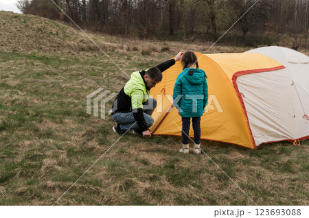Camping adventure unfolds as a child and an adult set up a bright orange tent on a grassy field surrounded by trees during a peaceful afternoon in early spring 123693088
