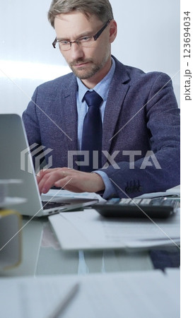 Businessman wearing blue suit is working with laptop, calculator and documents at the glass table in office set. Low lighting, vertical portrait. Business people and audit Businessman wearing blue suit is working with laptop, calculator and documents at the glass table in office set. Low lighting, vertical portrait. Business people and audit 123694034