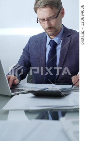 Businessman wearing blue suit is working with laptop, calculator and documents at the glass table in office set. Low lighting, vertical portrait. Business people and audit Businessman wearing blue suit is working with laptop, calculator and documents at the glass table in office set. Low lighting, vertical portrait. Business people and audit 123694036