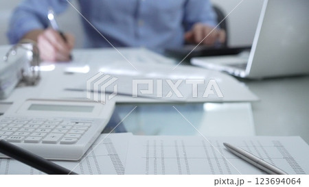 Calculator, pen and documents are lying on accountant's desk. Businessman wearing blue shirt is working with documents on the background. Business people, audit concept 123694064