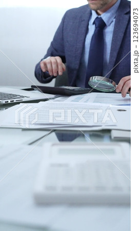 Businessman wearing blue suit is analyzing financial reports with magnifying glass and using calculator in a professional office setting. Low lighting, close-up vertical view. Business people, audit 123694073
