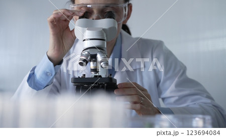 Woman scientist wearing lab coat and protective glasses is using microscope in modern laboratory setup. Low key lighting photo. Science and Medicine Woman scientist wearing lab coat and protective glasses is using microscope in modern laboratory setup. Low key lighting photo. Science and Medicine 123694084