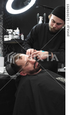 Professional barber carefully shaving client's beard with straight razor, applying gel and using hot towel in contemporary barbershop setting, vertical view Professional barber carefully shaving client's beard with straight razor, applying gel and using hot towel in contemporary barbershop setting, vertical view 123694093