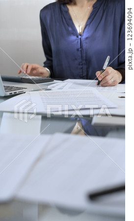 Businesswoman wearing blue dress is working with laptop, calculator and documents in a professional office setting. Low lighting, close-up vertical view. Business people, audit concept Businesswoman wearing blue dress is working with laptop, calculator and documents in a professional office setting. Low lighting, close-up vertical view. Business people, audit concept 123694094