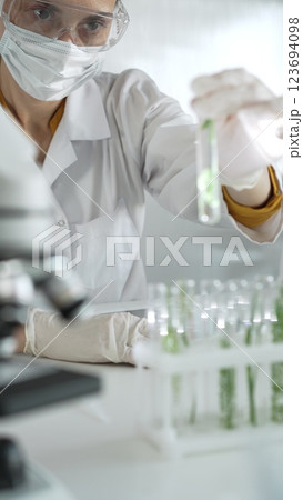 Woman scientist wearing lab coat, white gloves, face mask and protective glasses, is holding a test tube with plants inside near microscope in laboratory, vertical portrait view. Science and medicine 123694098