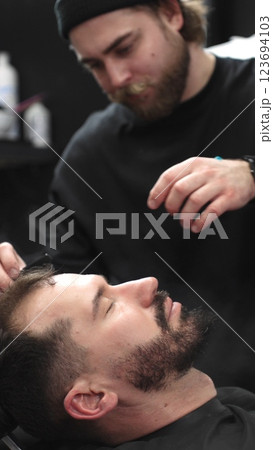 Professional barber carefully shaving client's beard with straight razor, applying gel and using hot towel for precise grooming in contemporary barbershop setting, vertical view 123694103