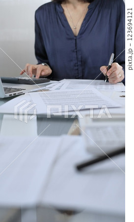 Businesswoman wearing blue dress is working with laptop, calculator and documents in a professional office setting. Low lighting, close-up vertical view. Business people, audit concept Businesswoman wearing blue dress is working with laptop, calculator and documents in a professional office setting. Low lighting, close-up vertical view. Business people, audit concept 123694121
