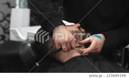 Professional barber carefully shaving client's beard with straight razor, applying gel and using hot towel for precise grooming in contemporary barbershop setting 123694149