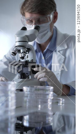 Man scientist wearing lab coat, white gloves, medical mask and protective glasses using microscope doing research in modern laboratory, portrait view. Low key lighting photo. Science and Medicine Man scientist wearing lab coat, white gloves, medical mask and protective glasses using microscope doing research in modern laboratory, portrait view. Low key lighting photo. Science and Medicine 123694150