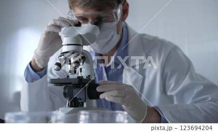 Man scientist researcher wearing white gloves, protective glasses and medical mask is using microscope in modern laboratory, portrait view. Low key lighting photo. Science and Medicine Man scientist researcher wearing white gloves, protective glasses and medical mask is using microscope in modern laboratory, portrait view. Low key lighting photo. Science and Medicine 123694204