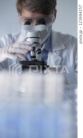 Man scientist wearing lab coat, white gloves, medical mask and protective glasses using microscope doing research in modern laboratory, portrait view. Low key lighting photo. Science and Medicine Man scientist wearing lab coat, white gloves, medical mask and protective glasses using microscope doing research in modern laboratory, portrait view. Low key lighting photo. Science and Medicine 123694257