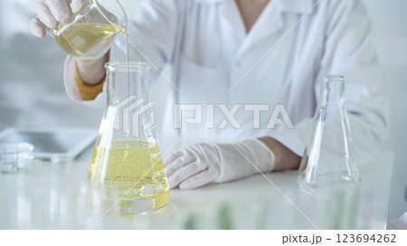 A scientist, wearing a lab coat and white protective gloves, is pouring a yellow oily liquid from one beaker to another in laboratory, close up. Medicine and science concept A scientist, wearing a lab coat and white protective gloves, is pouring a yellow oily liquid from one beaker to another in laboratory, close up. Medicine and science concept 123694262